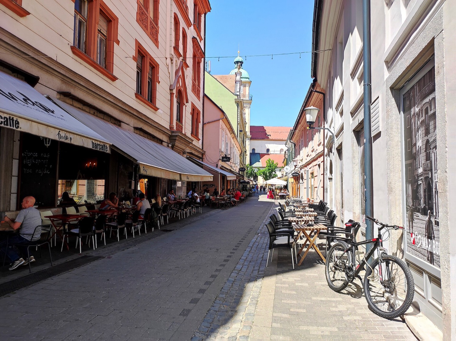 Cafe terraces along Poštna ulica in summer sun, people at the outdoor tables, the cathedral tower closing the view